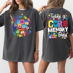 May include: Dark gray t-shirts with colorful graphics. One shirt features the text "It's okay to feel all the feels" with cartoon characters. The other shirt has "Today is a Core Memory Day" with cartoon characters.
