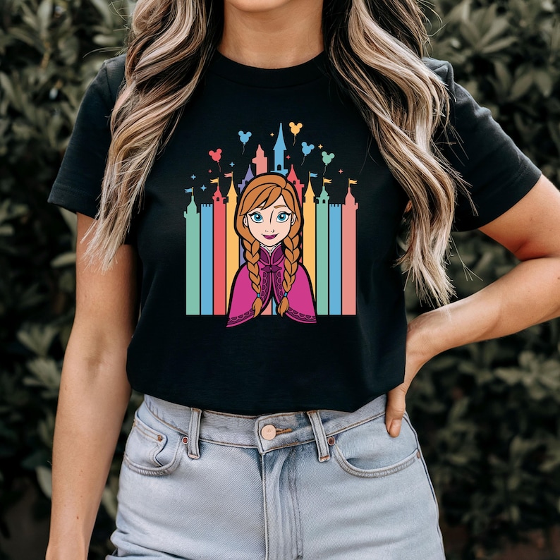 May include: Black t-shirt featuring a colourful graphic of Anna from Frozen in front of a rainbow castle with Mickey Mouse balloons.