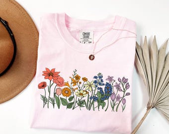 May include: A light pink t-shirt with a floral design across the bottom. The design features various colorful flowers, including red, orange, yellow, blue, and purple. The shirt has a Comfort Colors tag and a gold necklace.