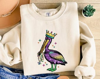 May include: A cream-colored sweatshirt with a purple, green, and gold glitter pelican wearing a crown and holding Mardi Gras beads. The pelican is on a white background with gold stars.