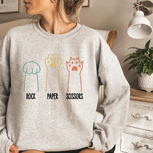 K&ouml;nnte beinhalten: Ein graues Sweatshirt mit einer Grafik von Katzenpfoten, die Stein, Papier, Schere spielen. Der Text "Stein, Papier, Schere" ist unter den Pfoten gedruckt.