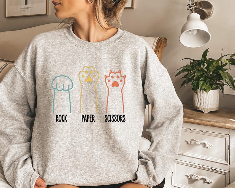 K&ouml;nnte beinhalten: Ein graues Sweatshirt mit einer Grafik von Katzenpfoten, die Stein, Papier, Schere spielen. Der Text "Stein, Papier, Schere" ist unter den Pfoten gedruckt.