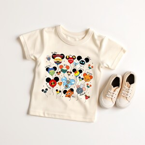 May include: Cream-colored toddler t-shirt featuring a colorful graphic of cartoon character balloons. The balloons are in the shape of Mickey Mouse heads and other characters. A pair of cream-colored canvas sneakers are next to the shirt.