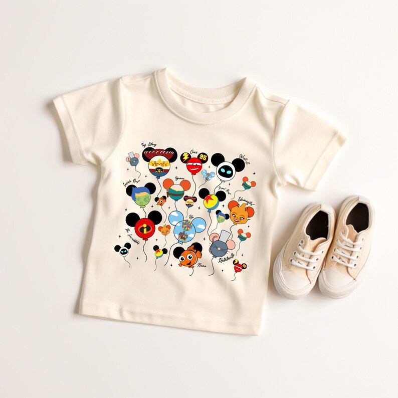 May include: Cream-colored toddler t-shirt featuring a colorful graphic of cartoon character balloons. The balloons are in the shape of Mickey Mouse heads and other characters. A pair of cream-colored canvas sneakers are next to the shirt.