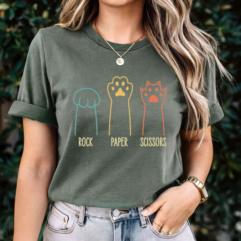 K&ouml;nnte beinhalten: Ein gr&uuml;nes T-Shirt mit einer Grafik von Katzenpfoten, die Stein, Papier, Schere spielen. Der Text "Stein, Papier, Schere" ist unter den Pfoten gedruckt.