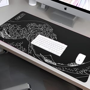 Puede incluir: Alfombrilla de escritorio rectangular negra con un contorno blanco de la Gran Ola de Hokusai. Un teclado y un ratón blancos están sobre la alfombrilla. La alfombrilla está sobre un escritorio blanco, con un monitor de ordenador al fondo.