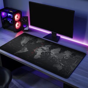 May include: A black and grey world map design mouse mat on a desk. The desk also has a computer monitor, speakers, and a computer tower with red and purple lights. The mouse mat is rectangular and covers a large portion of the desk.
