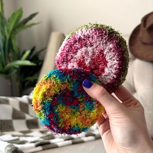 May include: Two round, textured scrubbers made of fuzzy yarn. One scrubber features a vibrant, multi-colored tie-dye pattern with shades of blue, yellow, orange, and pink. The other scrubber has a pink center with a green outer edge.