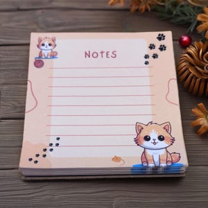 May include: A notepad with a light peach background featuring a cartoon cat illustration. The word "NOTES" is printed at the top. The notepad has lined pages and paw print and mouse illustrations.