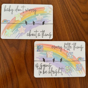 Pode incluir: Dois cartões com um fundo de arco-íris e o texto "baby don't worry about a thing" e "every little thing is going to be alright". Cada cartão tem três pássaros pretos em um fio e estrelas e quadrados brilhantes.