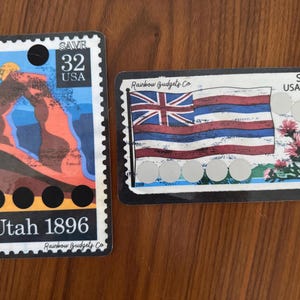 Op de afbeelding: Twee rechthoekige stickers die op postzegels lijken. Eén toont een Utah-landschap met de tekst "Utah 1896" en "SAVE 32 USA". De andere toont een vlagontwerp met de tekst "SAVE USA 42 HAWAII".