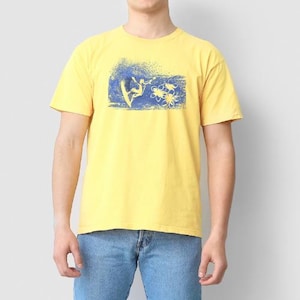 May include: A bright yellow short-sleeve t-shirt featuring a blue graphic print on the chest. The design depicts a surfer riding a wave alongside three stylized tropical flowers. The shirt has a crew neckline and a standard fit construction.