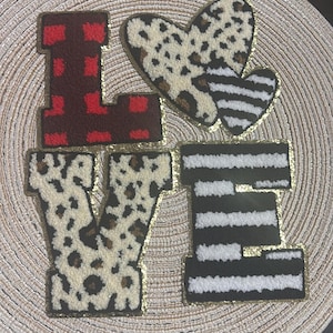 Puede incluir: Una pieza decorativa que deletrea "LOVE" con letras individuales y un corazón. Las letras tienen diferentes patrones: cuadros rojos, estampado de leopardo y rayas blancas y negras. Un borde dorado perfila cada elemento.