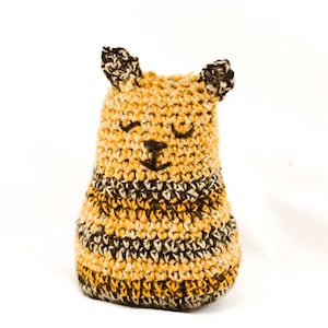 May include: A small, hand-crocheted plush toy shaped like a bee, featuring a rounded body with horizontal stripes in yellow, black, and white yarn. The toy has two small pointed ears at the top and embroidered black thread details for closed eyes and a nose.
