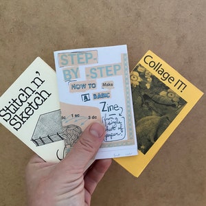 Op de afbeelding: Drie handgemaakte zines worden in een hand gehouden. De zines heten "Stitch n' Sketch", "Step-by-Step How to Make a Basic Zine" en "Collage IT!". De zines hebben verschillende kleuren, waaronder crème, geel en wit.