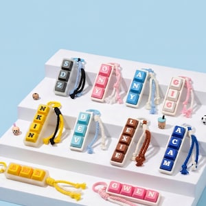 May include: A collection of colorful keychains, each featuring a name spelled out with individual letter blocks. The keychains have various color schemes, including yellow, pink, blue, and brown, with matching rope accents. The background is a light blue.
