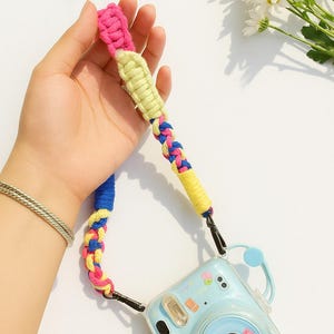 Puede incluir: Una cámara instantánea azul claro con una correa trenzada colorida. La correa presenta secciones rosas, amarillas y azules. La cámara tiene una funda transparente con pegatinas decorativas.