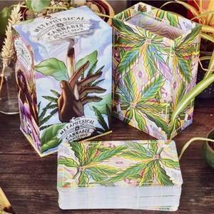 May include: The Metaphysical Cannabis Oracle Deck box is green, yellow, and blue with a cannabis leaf design. The deck of cards is stacked and features a similar design. The box has the title "The Metaphysical Cannabis Oracle Deck" printed on it.