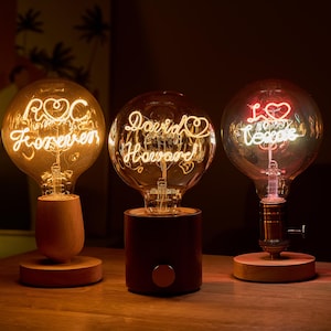 Puede incluir: Tres bombillas decorativas con mensajes personalizados. Las bombillas están sobre bases de madera y tienen una luz cálida e incandescente. Los mensajes en las bombillas son "ROC Forever", "David Howard" y "I Love You Too".
