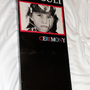 May include: A black and white album cover with the band name "THE CULT" in white text and the album title "CEREMONY" in red and blue text. The cover features a black and white photo of a young person with a painted face.