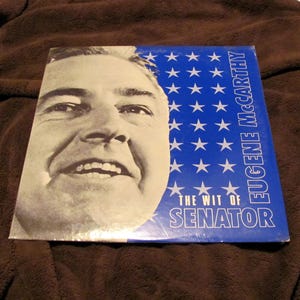 Könnte beinhalten: Vintage Vinyl-Schallplattenalbum mit einem Schwarz-Weiß-Porträt eines Mannes. Das Albumcover hat einen blauen Hintergrund mit weißen Sternen und dem Text "EUGENE MCCARTHY" und "THE WIT OF SENATOR".