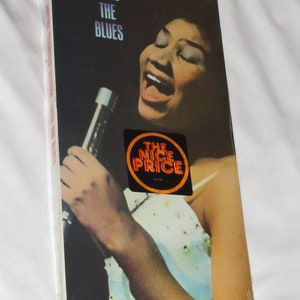 May include: A compact disc case with a photo of Aretha Franklin singing into a microphone. The case is black with white text that reads "Aretha Sings The Blues". The case also has a sticker that reads "The Nice Price".