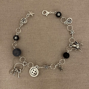 Puede incluir: Pulsera de abalorios plateada con una variedad de dijes, incluyendo estrellas, una ara&ntilde;a, un bot&oacute;n y llaves. La pulsera tambi&eacute;n incluye cuentas negras y grises, conectadas por peque&ntilde;os anillos plateados.