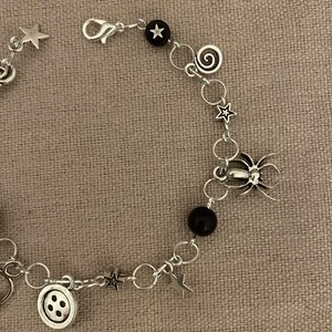 Puede incluir: Pulsera de abalorios plateada con estrellas, una ara&ntilde;a, un bot&oacute;n y espirales. La pulsera tambi&eacute;n incluye cuentas negras y un cierre. La pulsera se muestra sobre una superficie de color neutro. Joya &uacute;nica.