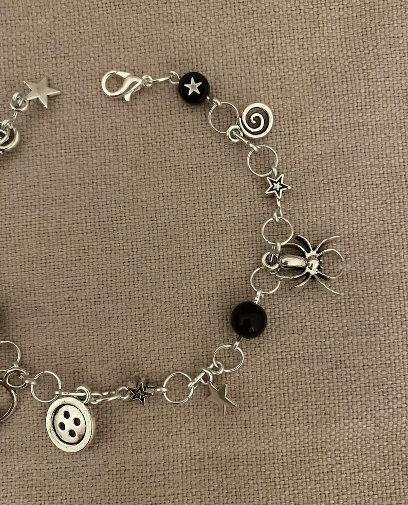 Puede incluir: Pulsera de abalorios plateada con estrellas, una ara&ntilde;a, un bot&oacute;n y espirales. La pulsera tambi&eacute;n incluye cuentas negras y un cierre. La pulsera se muestra sobre una superficie de color neutro. Joya &uacute;nica.