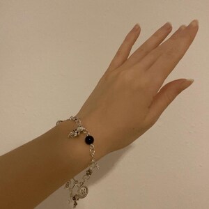 Puede incluir: Pulsera de abalorios plateada con una cuenta negra y varios abalorios, incluyendo estrellas y una carita sonriente. La pulsera se lleva en la mu&ntilde;eca sobre un fondo blanco liso. La pulsera es de metal color plata.