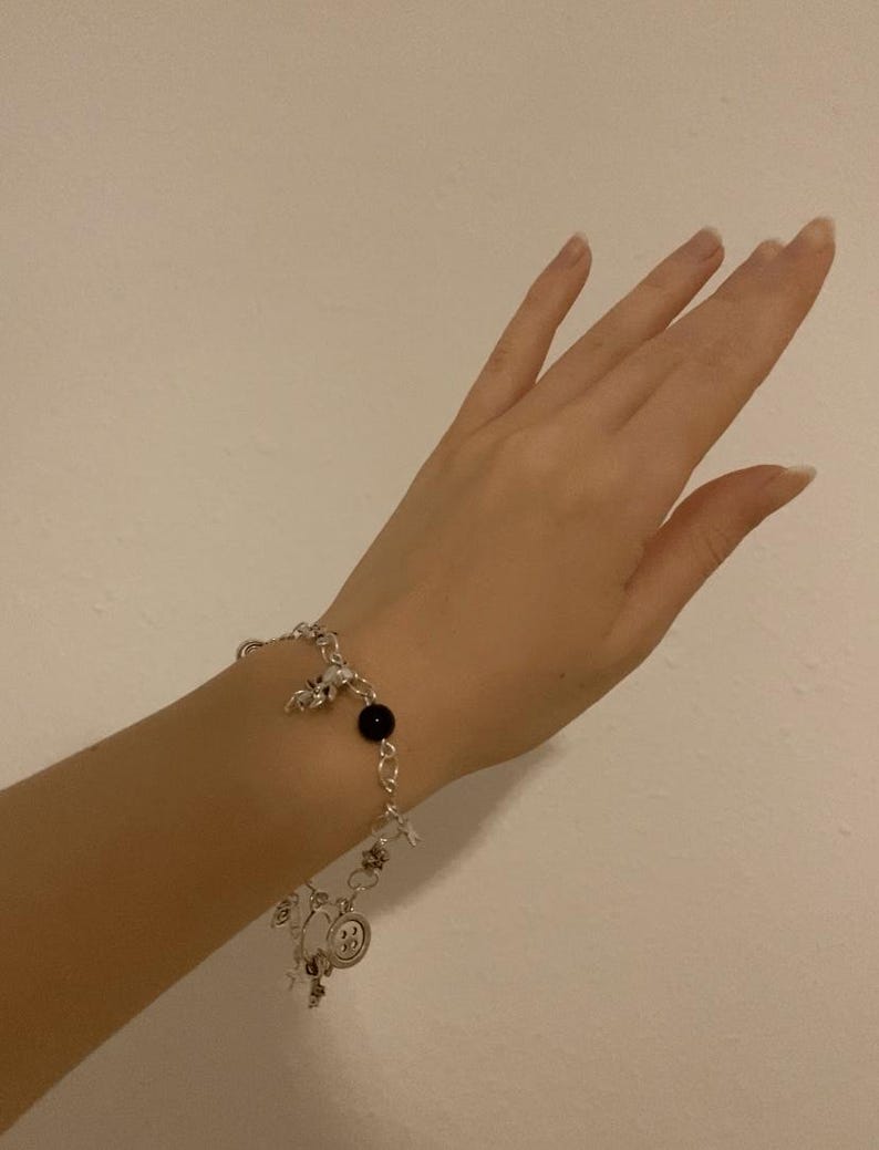 Puede incluir: Pulsera de abalorios plateada con una cuenta negra y varios abalorios, incluyendo estrellas y una carita sonriente. La pulsera se lleva en la mu&ntilde;eca sobre un fondo blanco liso. La pulsera es de metal color plata.