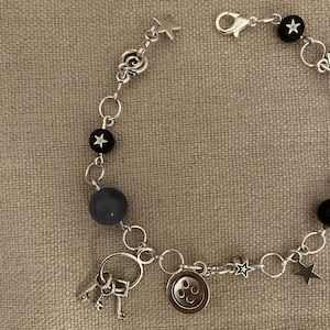 Puede incluir: Pulsera plateada con una mezcla de cuentas y dijes. Incluye cuentas negras con detalles de estrellas blancas, una cuenta gris y dijes en forma de estrella. Los dijes adicionales incluyen un bot&oacute;n y un juego de llaves.