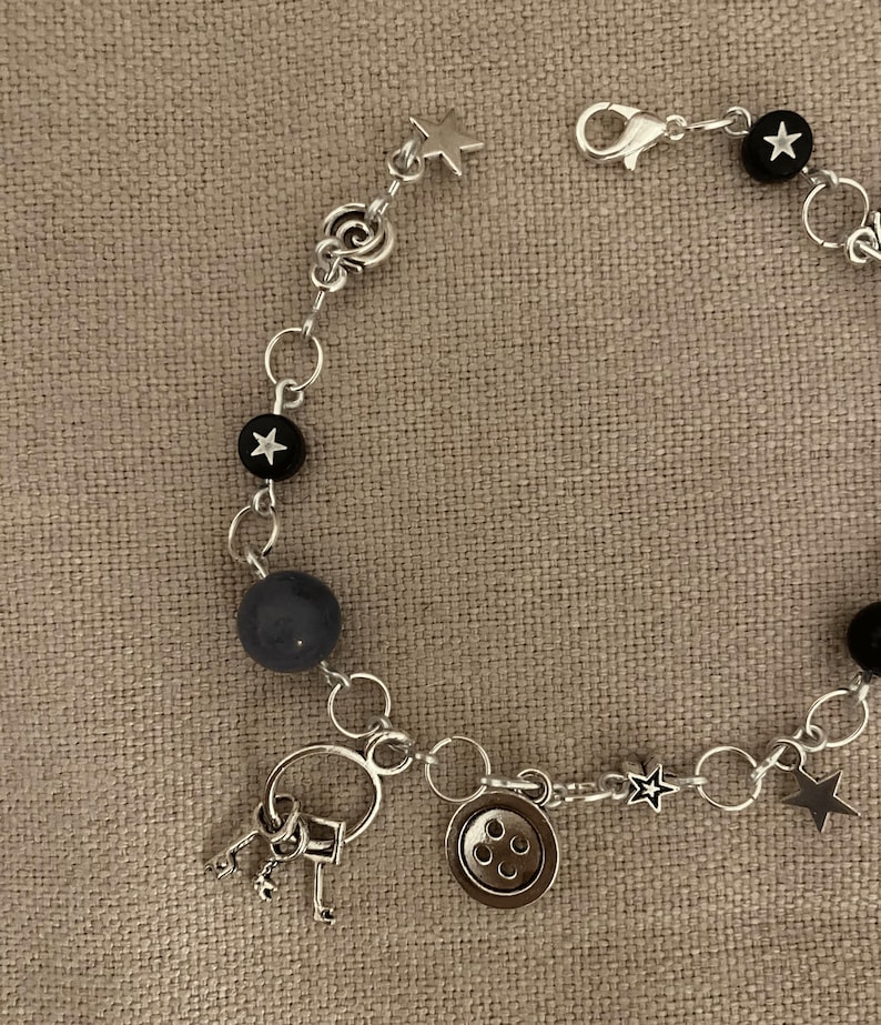 Puede incluir: Pulsera plateada con una mezcla de cuentas y dijes. Incluye cuentas negras con detalles de estrellas blancas, una cuenta gris y dijes en forma de estrella. Los dijes adicionales incluyen un bot&oacute;n y un juego de llaves.