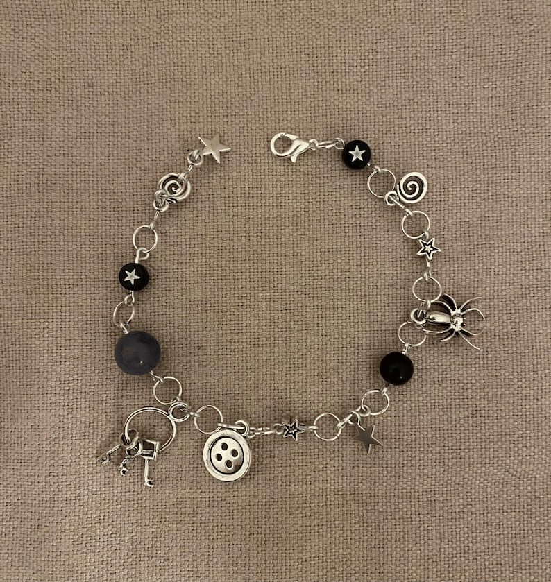 Puede incluir: Pulsera de abalorios plateada con una variedad de dijes, incluyendo estrellas, una ara&ntilde;a, un bot&oacute;n y llaves. La pulsera tambi&eacute;n incluye cuentas negras y grises, conectadas por peque&ntilde;os anillos plateados.