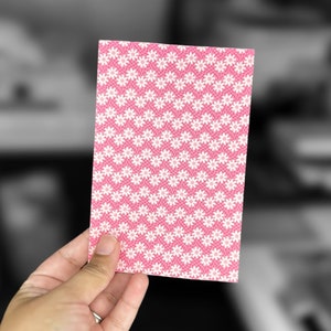 May include: A pink card with a white daisy pattern. The card is being held by a hand.
