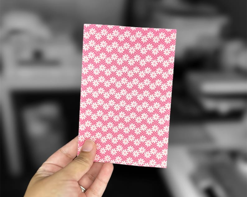 May include: A pink card with a white daisy pattern. The card is being held by a hand.