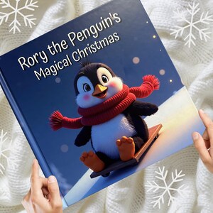 Può includere: Un libro per bambini intitolato "Rory the Penguin's Magical Christmas" presenta un pinguino dei cartoni animati con una sciarpa rossa, che va sullo slittino. La copertina ha uno sfondo blu con fiocchi di neve ed è tenuta da mani.