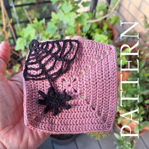 May include: A crocheted square in a dusty rose color, featuring a black spider and web design. The spider hangs from the web, which is attached to the top corner of the square. The word "PATTERN" is printed vertically on the right side.