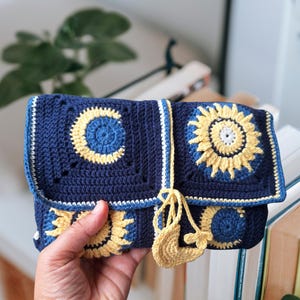 Può includere: Pochette all'uncinetto blu navy con disegni di girasoli e lune gialle e bianche. La pochette ha una chiusura a patta e un nappa gialla. La borsa è tenuta in mano da una persona.