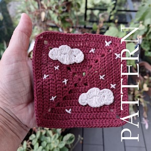 May include: A square, crocheted piece in a deep red hue, adorned with white cloud appliqués and small white cross-stitch accents. The word "PATTERN" is vertically displayed in white. The piece is held in a hand.