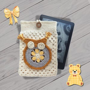 Könnte beinhalten: Cremefarbene gehäkelte Kindle-Hülle mit einem braunen Eulen-Design mit gelben und grauen Akzenten. Die Hülle hat einen Knopfverschluss und Quasten-Details. Ein Kindle E-Reader ist teilweise sichtbar.