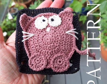 May include: A crocheted cat square with pink and white yarn. The cat has a black border and white eyes.