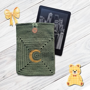 Puede incluir: Funda para lector electrónico de crochet verde con un botón marrón y una letra C amarilla en el centro.