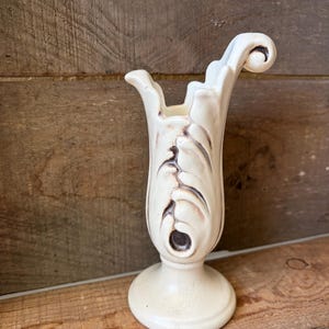 May include: A cream-colored ceramic vase featuring a pedestal base and a flared, asymmetrical rim with a curled top edge. The body of the vase is decorated with a raised, leaf-like relief pattern accented with dark brown shading in the crevices.