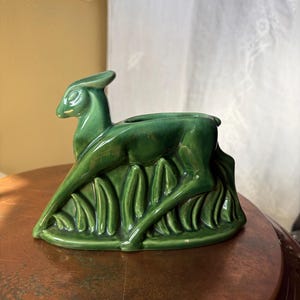 Puede incluir: Una maceta de cerámica de color verde oscuro brillante con forma de ciervo reclinado que descansa sobre un campo de hierba estilizada. El ciervo está representado en posición tumbada con la cabeza erguida y la abertura para plantas en el lomo.