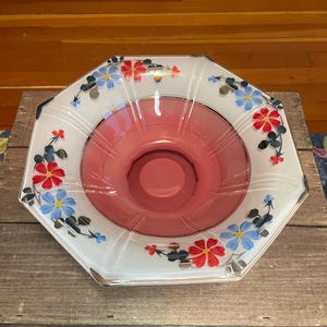 May include: An octagonal glass bowl with a white exterior and a pink interior. The rim is decorated with red and blue floral patterns. The bowl sits on a wooden surface.