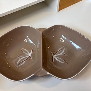 May include: A taupe-colored ceramic divided serving dish with a symmetrical, dual-bowl design. Each bowl features a painted graphic of white and dark red stylized leaves at the center. The dish has a smooth, glossy finish and a white rim.
