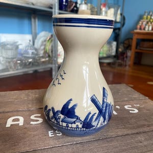 May include: A cream-coloured vase with blue windmill and bird illustrations. The vase has a flared top with two blue stripes and a bulbous body. The base of the vase has a blue border. The vase is on a wooden surface.