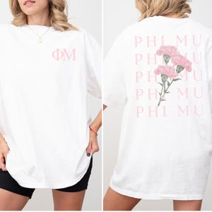 Op de afbeelding: Wit T-shirt met de Griekse letters &Phi;&Mu; in lichtroze op de voorkant. De achterkant van het shirt heeft een bloemenontwerp van roze anjers en de woorden PHI MU in roze.