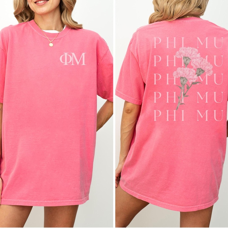 Op de afbeelding: Een koraalkleurig t-shirt met de Griekse letters &Phi;&Mu; op de voorkant. De achterkant van het shirt heeft de tekst "PHI MU" verticaal herhaald met een bloemenontwerp. Het shirt heeft een relaxte pasvorm en korte mouwen.